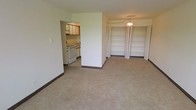 This single-floor unit presents an open, practical layout that combines the living area and kitchen in a cohesive, easy-to-navigate floor plan. The main living space is finished with neutral-toned carpet, white walls, and dark baseboards that frame the room cleanly. A pendant light fixture hangs near the storage wall, providing a subtle focal point overhead.

Two sets of built-in open shelving units with bifold doors occupy one wall, offering substantial storage capacity for books, linens, or personal items. Their white finish keeps the area feeling bright and organized.

The kitchen, visible through an open partition wall, features white cabinetry, a white range with oven, and a sink positioned beneath upper cabinets. The layout is compact yet functional, with flooring that transitions from the carpeted living area. The partial wall separating the kitchen creates a sense of definition between the two spaces while maintaining an open, connected feel.

Overall, the unit presents a straightforward, functional design with ample built-in storage, a well-integrated kitchen, and a clean neutral palette throughout.