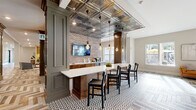 This thoughtfully designed single-floor amenity space masterfully combines industrial character with refined, classic detailing. The centerpiece is a sophisticated lounge area showcasing a striking herringbone-patterned wood floor that flows throughout much of the space, anchored by plush seating, a leather bench, and a navy blue paneled feature wall housing a mounted television. Natural light pours in through multiple windows, enhancing the warm, welcoming ambiance.

Adjacent to the lounge, a stylish bar and social area commands attention with an ornate pressed-tin ceiling, exposed brick accent walls, and Edison-style pendant lighting suspended overhead. A long white quartz-topped island with dark wood base provides generous bar seating, while built-in cabinetry and a mounted television complete the entertainment-ready setup. The floor transitions here to a bold geometric patterned tile, adding visual interest.

A games area offers a billiards setup alongside open shelving and paneled wainscoting in muted blue-gray tones. The space also includes a dedicated conference room with a light wood table, upholstered seating, a wall-mounted display, and frosted interior windows that maintain privacy while allowing natural light. Connecting corridors feature clean herringbone flooring and tasteful accent pieces, tying the entire space together seamlessly.