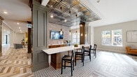 This thoughtfully designed single-floor amenity space masterfully combines industrial character with refined, classic detailing. The centerpiece is a sophisticated lounge area showcasing a striking herringbone-patterned wood floor that flows throughout much of the space, anchored by plush seating, a leather bench, and a navy blue paneled feature wall housing a mounted television. Natural light pours in through multiple windows, enhancing the warm, welcoming ambiance.

Adjacent to the lounge, a stylish bar and social area commands attention with an ornate pressed-tin ceiling, exposed brick accent walls, and Edison-style pendant lighting suspended overhead. A long white quartz-topped island with dark wood base provides generous bar seating, while built-in cabinetry and a mounted television complete the entertainment-ready setup. The floor transitions here to a bold geometric patterned tile, adding visual interest.

A games area offers a billiards setup alongside open shelving and paneled wainscoting in muted blue-gray tones. The space also includes a dedicated conference room with a light wood table, upholstered seating, a wall-mounted display, and frosted interior windows that maintain privacy while allowing natural light. Connecting corridors feature clean herringbone flooring and tasteful accent pieces, tying the entire space together seamlessly.