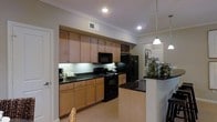 This single-floor kitchen is designed with both functionality and style in mind. The space is anchored by warm-toned wood cabinetry that extends nearly to the ceiling, providing generous storage throughout. Upper and lower cabinets line the main wall, offering ample organization for cookware, pantry items, and kitchen essentials, with open shelving above the refrigerator for additional display or storage.

The countertops are finished in dark granite, providing a rich contrast against the lighter cabinetry and creating a polished aesthetic. Black appliances, including a freestanding range with oven, built-in microwave, and a large refrigerator, complement the overall color scheme cohesively.

A peninsula with matching dark granite extends into the space, serving as both a food preparation area and a casual dining or entertaining surface. Pendant lighting hangs above the peninsula, adding focused illumination and a decorative element. Recessed lighting and under-cabinet lighting ensure the workspace is well-lit throughout.

Crown molding along the ceiling adds an architectural finishing touch, while light tile flooring grounds the space and enhances the overall sense of brightness and openness.