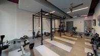 Located at 560 E Stonewall St in Charlotte's Uptown neighborhood, this single-floor fitness center is designed to accommodate a comprehensive range of workout routines. The space is organized into distinct functional zones, offering both cardio and strength training areas alongside a dedicated free weights and functional training section.

The interior design features a neutral color palette accented by deep burgundy walls and exposed structural beams painted to match, lending an urban, modern aesthetic. Linear LED lighting runs along the ceiling, complementing the abundant natural light that streams in through large windows lining multiple walls. These windows also provide views of the surrounding streetscape, creating an open, connected atmosphere.

The flooring throughout consists of a distinctive herringbone-patterned tile in alternating neutral tones, adding a polished, contemporary look. The cardio zone is outfitted with treadmills, ellipticals, and stationary bikes positioned near the windows. Rows of selectorized strength machines occupy the central and secondary areas, while the free weights section features multi-station power racks, barbells, weight plates, and adjustable benches. Ceiling fans are distributed throughout to maintain air circulation. Wall-mounted televisions and dual drinking fountains set against a dark tile backsplash complete the amenities, ensuring a fully functional fitness environment.