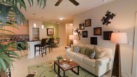 This single-floor space showcases an open-concept layout that seamlessly connects the living room, dining area, and kitchen into one cohesive, flowing environment. The living area is anchored by a comfortable cream sofa adorned with nature-inspired throw pillows, complemented by a decorative green area rug featuring a floral motif. A wooden coffee table set serves as a functional centerpiece. The walls are adorned with an eclectic gallery of dark-framed artwork alongside striking decorative gecko sculptures, lending the space a distinctive tropical personality. Pendant lighting and a ceiling fan overhead provide both practical illumination and casual comfort. The kitchen and dining area are separated by a breakfast bar with bar-height seating, while the dining space accommodates a full table and chairs beneath track lighting. An accent wall in soft green adds depth and visual interest to the kitchen zone. Neutral tile flooring runs throughout, tying the spaces together cohesively. Overall, the design blends relaxed, tropical aesthetics with functional open-plan living, creating a bright and inviting atmosphere.