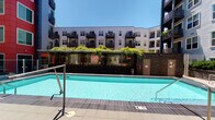 This single-floor space is associated with a multi-story residential complex that features a well-maintained outdoor pool area. The pool is generously sized, framed by a tiled surround and a pergola draped in lush greenery, creating a resort-like atmosphere. The surrounding building facade showcases a modern design with private balconies and dark metal railings set against a clean white and gray exterior.

The interior includes a bathroom finished in warm, neutral tones. The vanity features an oak wood cabinet base topped with a light countertop and an integrated sink, paired with classic chrome dual-handle faucets. A framed mirror with a wood-toned border is mounted above, complemented by a multi-bulb vanity light fixture that provides ample illumination. A metal over-the-toilet shelving unit offers practical storage space. Interior doors are finished in white with brass round knob hardware, and electrical outlets feature matching brass cover plates, giving the space a consistent, classic aesthetic. The walls throughout are painted in soft, neutral tones, contributing to a clean and understated ambiance.