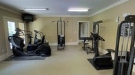 This single-floor fitness space is thoughtfully laid out to accommodate a range of exercise needs. The room features neutral beige walls, white baseboards, and a durable speckled rubberized floor surface that provides both safety and easy maintenance. Overhead fluorescent lighting ensures the space is well-illuminated throughout.

The room is furnished with a selection of fitness equipment, including two elliptical cross-trainers positioned on either side of the room, a cable pulley lat pulldown machine centrally located, and a weight bench with a rack toward the rear. Additional strength training equipment is also present, offering options for a variety of muscle groups. A wall-mounted television is installed near the entrance, allowing users to watch content during cardio sessions.

A doorway at the rear of the room leads to an adjacent space, suggesting additional amenities may be accessible nearby. The overall layout is open and organized, allowing users to move freely between equipment. The space conveys a practical, no-frills atmosphere focused entirely on functionality, making it well-suited as a dedicated fitness facility for regular use.