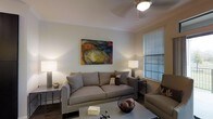 This single-floor residence offers a thoughtfully designed living space that balances comfort and contemporary style. The living room serves as the centerpiece of the home, featuring a generously sized neutral-toned sofa with nailhead trim detailing, complemented by a matching armchair that creates an inviting seating arrangement. A sleek coffee table anchors the space, while coordinating end tables with elegant floor lamps provide warm, layered ambient lighting throughout the room. A bold, colorful abstract painting mounted on the wall adds a striking artistic focal point, infusing the space with energy and personality. Crown molding along the ceiling lends a refined architectural touch, while a ceiling fan ensures year-round comfort. Dark hardwood flooring grounds the room with a rich, classic aesthetic. A large window dressed with horizontal blinds allows natural light to filter in, brightening the interior while maintaining privacy. The overall ambiance of this residence is warm and inviting, with a cohesive design palette that blends neutral tones with carefully chosen accents, creating a space that feels both livable and polished.