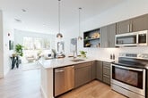 This well-appointed single-floor residence at 128 Maple St in Danvers, MA offers a thoughtfully designed open-concept layout across three bedrooms and two full bathrooms. The modern kitchen is a standout feature, showcasing grey cabinetry, a white quartz waterfall island with bar seating, stainless steel appliances including a dishwasher and range, a subway tile backsplash, and sleek globe pendant lighting. The kitchen flows seamlessly into the living room, which is bathed in natural light from oversized windows offering street-level views of the surrounding neighborhood. Light wood-tone flooring runs throughout, lending warmth and continuity to the entire space. The primary bedroom is generously sized with a large window and dedicated closet space, while additional bedrooms are bright and comfortable with ample room. Both bathrooms are clean and functional, with one featuring tile flooring and a tub-shower combination with classic fixtures. A dedicated laundry room houses a stacked washer and dryer unit alongside additional shelving. Two closets provide convenient storage throughout. The overall aesthetic is contemporary and airy, with white walls, clean lines, and abundant natural light creating a bright, inviting atmosphere throughout.