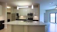 This single-floor residence showcases a thoughtfully designed open-concept layout that maximizes both functionality and visual appeal. The kitchen serves as a central highlight, featuring a striking two-tone design with crisp white upper cabinetry and dark lower cabinets, creating a bold and modern contrast. A generous granite-topped island anchors the space, offering ample room for meal preparation and casual dining, complete with an undermount stainless steel sink. Stainless steel appliances, including a French door refrigerator, electric range, and built-in microwave, lend a polished, professional quality to the kitchen. A white subway tile backsplash adds a clean, classic touch that complements the contemporary aesthetic. Three pendant lights hang above the kitchen area — two cylindrical white pendants and a linear fluorescent fixture — providing focused task lighting while enhancing the room's modern character. The open sightlines from the kitchen connect seamlessly to adjacent living areas, promoting an airy, connected atmosphere throughout the home. Neutral wall tones and consistent flooring tie the spaces together cohesively, creating a bright and welcoming environment throughout this well-appointed residence.