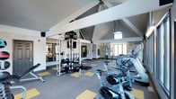 This single-floor fitness center is thoughtfully designed to accommodate a wide range of workout needs. The space is immediately distinguished by its dramatic vaulted ceiling with exposed white beams, which creates an open, airy atmosphere and adds architectural character to the room. A ceiling fan is centrally mounted to maintain airflow throughout the space during workouts.

The flooring features a distinctive patterned carpet in gray and yellow, adding a vibrant, energetic feel that complements the facility's active purpose. Natural light filters in through multiple windows, brightening the interior and enhancing the overall workout environment.

The facility is comprehensively outfitted with a row of stationary cycling bikes and additional cardio equipment positioned near the windows, allowing users to exercise while enjoying natural light. A multi-station strength training machine, along with free weights and various accessories, occupies the central area of the floor. Motivational typography wall art featuring fitness-related words adds an inspiring visual element to the space.

Large mirrors line one wall, expanding the sense of space and allowing users to monitor their form during exercise. The facility appears to connect to an adjacent area, visible through glass doors, potentially offering additional workout or functional training space. Overall, the fitness center presents a modern, well-appointed environment suited for a full-body workout routine.