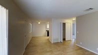 This single-floor residence offers a clean and functional layout designed for comfortable everyday living. The main living area is finished with light-toned hardwood flooring that extends throughout, lending warmth and continuity to the space. A textured popcorn ceiling with a centrally mounted ceiling fan overhead provides both airflow and a classic aesthetic touch. Neutral white walls keep the interior bright and versatile, ready to accommodate a variety of décor styles.

Natural light filters in through a window along the main wall, contributing to an airy and open atmosphere. A floor-level HVAC vent indicates a central climate control system servicing the unit. Toward the rear of the living area, a pass-through view reveals a kitchen equipped with dark appliances and cabinetry, set against a lighter backdrop.

A central hallway branching off the main room provides access to additional areas of the home. Flanking the hallway are bifold closet doors offering generous built-in storage, while a doorway leads further into what appears to be a carpeted room, adding a softer surface underfoot in that area. The overall layout is straightforward and well-organized, making efficient use of the available space across its single level.