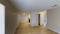 This single-floor residence offers a clean and functional layout designed for comfortable everyday living. The main living area is finished with light-toned hardwood flooring that extends throughout, lending warmth and continuity to the space. A textured popcorn ceiling with a centrally mounted ceiling fan overhead provides both airflow and a classic aesthetic touch. Neutral white walls keep the interior bright and versatile, ready to accommodate a variety of décor styles.

Natural light filters in through a window along the main wall, contributing to an airy and open atmosphere. A floor-level HVAC vent indicates a central climate control system servicing the unit. Toward the rear of the living area, a pass-through view reveals a kitchen equipped with dark appliances and cabinetry, set against a lighter backdrop.

A central hallway branching off the main room provides access to additional areas of the home. Flanking the hallway are bifold closet doors offering generous built-in storage, while a doorway leads further into what appears to be a carpeted room, adding a softer surface underfoot in that area. The overall layout is straightforward and well-organized, making efficient use of the available space across its single level.