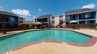 This outdoor pool area serves as a central amenity, offering residents a refreshing retreat within a secure, iron-fenced enclosure. The pool itself is a generously sized, kidney-shaped basin with clear turquoise water, framed by a classic red brick coping and a spacious concrete deck. The deck is furnished with woven lounge chairs ideal for sunbathing, as well as shaded seating areas featuring bright umbrella-topped tables, providing options for both sun and shade throughout the day. Tall fan palms and established shrubs add natural greenery and a relaxed, tropical character to the space. Access to the pool area is provided via a concrete walkway flanked by the iron perimeter fence, ensuring a safe and defined entry point. A posted pool rules sign and emergency phone signage reflect the safety-conscious design of the space. The surrounding two-story residential buildings, finished in a combination of brick and bold blue and brown exterior accents with pitched rooflines, create an intimate, courtyard-like atmosphere. Under open skies, the pool area benefits from abundant natural light, making it a vibrant and inviting outdoor space suited for relaxation and recreation.