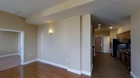 This single-floor space offers a clean, modern layout defined by a warm and neutral aesthetic. The interior is finished with rich hardwood flooring that extends through the main living area, transitioning to tile near the entryway, creating a natural visual distinction between zones. Smooth, taupe-toned walls are complemented by crisp white baseboards and trim, lending the space a polished, cohesive look throughout. A partial wall divides the main living area, adding architectural interest while maintaining an open feel. Recessed ceiling lights illuminate the hallway leading to the entry, while a wall-mounted sconce in the living area adds a warm, ambient glow. The entryway features a solid wood front door flanked by a secondary white door, providing both character and functionality. The ceiling design incorporates subtle tray-like architectural detailing, adding depth to the overall layout. The neutral palette and thoughtful lighting make this space versatile and move-in ready, well-suited for a variety of furnishing styles. Overall, the space conveys a sense of understated comfort and practical elegance across its single level.