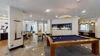 This single-floor amenity space presents a refined, contemporary design characterized by soaring double-height ceilings and floor-to-ceiling glass walls that flood the interior with natural light and frame views of lush palm-lined outdoor areas. Polished marble-look tile flooring flows throughout the main areas, creating a seamless, upscale aesthetic. The grand entrance lobby features sculptural seating, including a striking woven lounge chair, vibrant blue-and-gold patterned area rugs, and a large wall-mounted display screen, establishing an immediate sense of arrival and sophistication.

A dedicated game and social lounge is anchored by a full-size billiards table beneath an eye-catching gold sculptural pendant light, complemented by a built-in illuminated aquarium wall and high-top marble dining tables with navy and cream seating. Adjacent to this area, a more intimate lounge zone offers curved banquette seating with layered accent pillows against a textured feature wall.

Glass-partitioned private meeting or conference rooms provide flexible gathering options, each furnished with wooden tables, media screens, and wall art. Throughout, the design palette of navy, gold, and warm neutrals creates a cohesive, resort-inspired ambiance. French doors in the dining area open directly to an outdoor pool courtyard, further enhancing the indoor-outdoor connection. Polished corridors with marble flooring lead to elevator access points, completing this well-appointed communal facility.