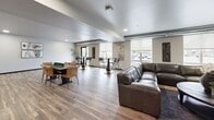 Located at 801 Kraewood Dr in Northfield, MN, this well-appointed clubroom serves as a premier community gathering space. The open-concept layout spans a generous single floor, accommodating multiple functional zones with ease. Wood-look plank flooring runs throughout, tying the space together with a warm, cohesive aesthetic.

The kitchen area is a standout feature, equipped with dark espresso cabinetry, granite-topped counters, a stainless steel refrigerator, and a large center island with an integrated sink and bar seating for multiple guests — ideal for catering or casual entertaining.

Adjacent to the kitchen, a spacious lounge area offers comfortable leather seating arranged around a patterned accent wall with a striking marbled wallcovering in cool blue and gold tones. Decorative cabinetry and potted greenery add warmth and character to this relaxed setting.

The dining and social zone is illuminated by natural light from several large windows overlooking the outdoors. This area features a mix of standard dining tables with leather chairs and tall bistro tables with bar stools, accommodating both casual gatherings and organized events.

Recessed lighting throughout ensures bright, even illumination, while the neutral walls and modern furnishings create a clean, contemporary ambiance suited for a wide range of community activities.