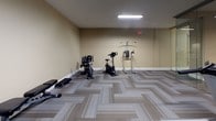 Located at 300 Canal in Lawrence, Massachusetts, this single-floor amenity space is designed to support both fitness and recreation. The layout is thoughtfully divided using floor-to-ceiling glass partitions, creating distinct yet visually connected zones while maintaining an open, airy feel throughout.

The fitness area is well-equipped with a range of cardio machines, including Spirit-brand treadmills, elliptical trainers, and stationary bikes. Strength and functional training options include a cable machine, a power tower, a rowing machine, and a dumbbell rack. Multiple wall-mounted flat-screen televisions are positioned throughout the workout zone, enhancing the exercise experience.

Adjacent to the fitness area, visible through the glass partition, is a recreation space featuring a billiards table and seating, offering a comfortable social alternative. The flooring throughout is a modern patterned carpet tile in neutral gray and taupe tones, providing both durability and visual cohesion. Recessed and fluorescent lighting illuminates the space evenly. The warm beige and slate-blue wall tones contribute to a clean, welcoming atmosphere suited for daily use.