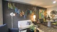 This single-floor space makes a strong visual impression through its thoughtfully designed living area. The focal point of the room is a dramatic dark-painted brick accent wall adorned with a cascading, handcrafted installation of multicolored wooden rods in shades of blue, green, and teal, adding depth and artistic character to the space. A vibrant abstract canvas painting featuring warm greens, oranges, and yellows is displayed alongside the installation, reinforcing the room's eclectic, gallery-like atmosphere. The living area is furnished with a tufted white sofa accented with patterned throw pillows, complemented by a round dark coffee table and textured drum-style side tables. A plush cream area rug grounds the seating arrangement, while a potted plant introduces a natural element. Industrial-style wall sconces provide warm, ambient lighting alongside a classic table lamp, creating a layered and inviting glow throughout the space. Crown molding along the ceiling adds a refined architectural detail that balances the room's edgier, artistic elements. Overall, the space blends contemporary design with bold, expressive décor choices.