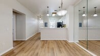 This single-floor residence is designed with a clean, open layout that promotes a bright and airy atmosphere throughout. Warm-toned wood-look flooring flows consistently across all living areas, tying the space together cohesively.

The kitchen is positioned centrally and features a peninsula with a light-colored countertop that serves as a natural divider between the kitchen and the open living area. White raised-panel cabinetry provides ample storage, while stainless steel appliances — including a refrigerator, electric range, and over-the-range microwave — lend a modern, functional aesthetic. A trio of pendant lights hangs above, adding both task lighting and visual interest.

The living area is generously proportioned and benefits from natural light filtering through a front-facing window with a transom detail. Ceiling fans are present in multiple rooms, enhancing comfort and airflow.

The bedrooms are well-sized, each featuring wood-look flooring, ceiling fans or ample natural light from large windows with outdoor greenery views, and closet storage. One bedroom includes mirrored sliding closet doors.

The bathroom is outfitted with a pedestal sink, a mirrored medicine cabinet with overhead lighting, and a shower-tub combination. A dedicated laundry room houses a full-size washer and dryer alongside wire shelving and a water heater.