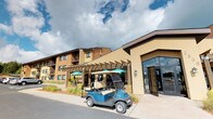 This unit is part of a well-established residential community defined by a warm blend of brick and stucco exterior finishes in earthy tan and brown tones. The building's facade is complemented by a prominent pergola structure near the main entrance, providing shaded outdoor seating beneath teal umbrella-covered tables — an inviting communal gathering space. Large floor-to-ceiling glass panels flank the entrance, allowing natural light to flow into the building's common areas. A golf cart parked near the entrance highlights the community's emphasis on accessible, convenient transportation throughout the property.

The surrounding grounds feature manicured landscaping with colorful plantings and maintained pathways. Ample parking is available nearby, with well-organized lots set against open sky views. Interior features visible include a closet space outfitted with wire shelving and hanging rods, offering practical storage solutions. Window treatments include horizontal slatted blinds throughout, providing adjustable light control and privacy.

The single-floor layout offers straightforward accessibility, making it a practical and functional living space within a larger community setting that balances outdoor amenities with comfortable interior design.