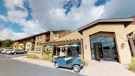 This unit is part of a well-established residential community defined by a warm blend of brick and stucco exterior finishes in earthy tan and brown tones. The building's facade is complemented by a prominent pergola structure near the main entrance, providing shaded outdoor seating beneath teal umbrella-covered tables — an inviting communal gathering space. Large floor-to-ceiling glass panels flank the entrance, allowing natural light to flow into the building's common areas. A golf cart parked near the entrance highlights the community's emphasis on accessible, convenient transportation throughout the property.

The surrounding grounds feature manicured landscaping with colorful plantings and maintained pathways. Ample parking is available nearby, with well-organized lots set against open sky views. Interior features visible include a closet space outfitted with wire shelving and hanging rods, offering practical storage solutions. Window treatments include horizontal slatted blinds throughout, providing adjustable light control and privacy.

The single-floor layout offers straightforward accessibility, making it a practical and functional living space within a larger community setting that balances outdoor amenities with comfortable interior design.