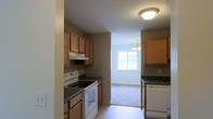 This single-floor residence offers a practical and inviting layout designed for comfortable everyday living. The kitchen is a standout feature, showcasing warm honey-toned wood cabinetry that provides ample storage space both above and below the countertops. Dark laminate counters offer a contrasting, easy-to-maintain work surface, while white appliances — including an electric range with overhead ventilation and a dishwasher — complete the functional setup. Tile flooring runs throughout the kitchen, providing a clean and durable foundation underfoot. A ventilation hood is positioned above the range, adding to the kitchen's practical design. The kitchen opens directly into an adjacent living area, which features plush carpeting and is bathed in natural light streaming through a large window. A pendant light fixture overhead adds a touch of warmth to this connected space. The neutral wall tones throughout create a calm, cohesive atmosphere that makes the home feel open and welcoming. The seamless transition between the kitchen and living area enhances the overall sense of flow, making the space well-suited for both daily routines and casual gatherings.