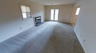 This two-story space offers a well-proportioned layout designed for comfortable living. The main living area is a standout feature, finished with plush wall-to-wall carpeting in a neutral gray tone that adds warmth underfoot. A classic brick fireplace serves as a natural focal point along one wall, providing both a functional heating element and a timeless architectural detail. Natural light fills the room through a side window fitted with a blind, while a set of French doors along the back wall introduces additional brightness and offers direct access to the outdoors. The white-painted walls and clean lines throughout contribute to a bright, open ambiance that feels move-in ready. The neutral palette throughout the living space creates a versatile backdrop suitable for a range of interior styles. With two floors, the overall layout provides separation between living and private spaces, lending the home a functional and organized flow. The ground floor living area, in particular, feels generously sized and airy, making it well-suited for both everyday relaxation and entertaining.