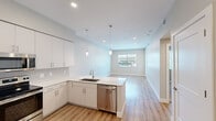 Located at 15024 N 90th St in Scottsdale, Arizona, this thoughtfully designed one-bedroom, one-bathroom residence offers a clean, modern aesthetic throughout its single-floor layout. The open-concept kitchen and living area serves as the heart of the home, featuring crisp white cabinetry that extends to the ceiling, sleek quartz countertops, and a full suite of stainless steel appliances including a French door refrigerator, electric range, microwave, and dishwasher. Pendant lighting accents the kitchen peninsula, which flows seamlessly into the living area. Light wood-look flooring runs throughout the main living spaces, complementing the neutral gray tones on the walls and creating a bright, airy atmosphere. The living room receives natural light through a large window and provides direct access to an outdoor area via a glass-panel door. The bedroom offers plush carpeting, a ceiling fan, and a window with outdoor views, providing a comfortable and restful retreat. The bathroom is well-appointed with a white vanity, walk-in glass shower, and consistent wood-look flooring. A dedicated laundry space houses a full-size stacked washer and dryer unit. The hallway connects all rooms efficiently, with clean white panel doors and ample closet storage completing this well-designed Scottsdale residence.