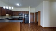 This single-floor unit offers a well-designed, open-concept layout that seamlessly connects the kitchen and living area, creating a spacious and functional living environment. Rich, dark hardwood flooring flows throughout, adding warmth and a cohesive aesthetic to the space.

The kitchen is a standout feature, equipped with rich dark wood cabinetry offering generous storage, complemented by dark countertops and a tasteful tile backsplash. Black appliances — including a refrigerator, gas range, and built-in microwave — provide a sleek, modern look. A peninsula with a breakfast bar overhang extends into the open living area, making it ideal for casual dining or entertaining. A pendant light fixture above the peninsula adds a refined touch to the kitchen zone.

The living area is bright and open, with neutral white walls that maximize the sense of space. A ceiling fan with a light kit provides both comfort and illumination. The layout flows naturally toward a hallway leading to additional rooms, with doors visible at the rear suggesting access to bedrooms and a closet or storage area. The clean lines, consistent flooring, and thoughtful design elements come together to create a comfortable and inviting single-floor living space.