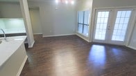 This single-floor space showcases a thoughtfully designed open living area that combines classic architectural details with a clean, modern aesthetic. The rich, dark hardwood flooring runs continuously throughout, providing a warm and polished foundation that anchors the entire space. Crown molding lines the ceiling perimeter, adding a refined architectural touch that complements the neutral gray-toned walls and bright white trim work.

A standout feature of the living area is the set of French doors, which flood the space with natural light and provide access to the outdoors. Flanking the French doors is a large window fitted with horizontal blinds, further enhancing the brightness of the interior. The combination of multiple windows and glass-paneled doors ensures the living area feels open and airy throughout the day.

The layout of the living space is generously proportioned, offering flexibility for various furniture arrangements. A partial white wall or built-in element is visible in the foreground, suggesting a natural division between the living area and an adjacent space such as a kitchen or entryway. Archways and open passages connect the rooms seamlessly, contributing to the overall flow and spaciousness of this well-appointed single-floor home.