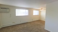 This single-floor space features an open living area designed with functionality and comfort in mind. The room is finished with wall-to-wall carpet in a neutral tan tone, providing a warm and understated foundation throughout. Two windows fitted with horizontal blinds are positioned along the main wall, allowing a generous amount of natural light to fill the space while offering adjustable privacy. The white walls and light-colored ceiling contribute to a clean, bright ambiance. A ceiling fan with an integrated light fixture is centrally mounted, offering both air circulation and overhead lighting. Baseboard heating units run along the base of the exterior walls, providing efficient climate control throughout the colder months. The layout is open and unobstructed, offering flexibility for a variety of furniture arrangements. Overall, the space presents a straightforward, practical interior with a neutral palette that serves as a blank canvas for personalization.