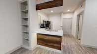 This single-floor space showcases a thoughtfully designed interior that blends modern aesthetics with warm, rustic accents. The kitchen serves as a central highlight, featuring grey shaker-style cabinetry, crisp white quartz countertops, and a full suite of stainless steel appliances including a gas range, microwave, dishwasher, and refrigerator. A peninsula with a rich wood-paneled base and an integrated sink anchors the kitchen, while a matching reclaimed wood beam overhead adds a distinctive architectural touch. Subway tile backsplash and recessed lighting complete the polished look. The open layout connects the kitchen seamlessly to the living area, which benefits from large windows fitted with white blinds that allow ample natural light to fill the space. Light wood-tone luxury vinyl plank flooring runs consistently throughout, lending warmth and cohesion to every room. Built-in open shelving near the entry provides practical display and storage space. The bathroom is clean and functional, equipped with a grey vanity cabinet, white countertop, a large mirror, and wood-look flooring that mirrors the rest of the unit. A front entry door with a paneled design completes the welcoming, move-in-ready feel of this well-maintained space.