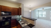 Located in Houston, TX 77070, this single-story residence offers a thoughtfully designed 1-bedroom, 2-bathroom layout that maximizes both comfort and functionality. The kitchen is a standout feature, showcasing rich dark wood cabinetry, dark granite countertops, a diamond-pattern tile backsplash, and black appliances including a range, microwave, and refrigerator. A double stainless steel sink sits at a peninsula that opens to the adjacent living and dining area via an elegant arched passageway framed by a decorative column, creating a seamless flow between spaces. The living area is generously sized, featuring plush carpeting, a ceiling fan, and multiple windows with blinds that invite ample natural light. A front entry door with a transom window further enhances the brightness of the main living space. The bedroom is well-proportioned, carpeted, and equipped with a ceiling fan and a window offering outdoor views. A second flexible room with carpet and a closet provides additional utility. Both bathrooms include tub-shower combinations with white tile surrounds and warm wood cabinetry. Tile flooring in the bathrooms and wood-look flooring in the kitchen complete the practical and cohesive interior design of this Houston residence.