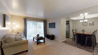 This single-floor residence offers an open and functional layout that connects the living, dining, and kitchen areas in a cohesive flow. The living room is carpeted and features a large sliding glass door dressed with vertical blinds and dark curtain panels, allowing generous natural light to fill the space while providing access to an outdoor area. A wall-mounted flat-screen television and a dark wood media console anchor the entertainment area, complemented by a comfortable sofa and a dark wood coffee table set atop a neutral area rug.

Transitioning from the living space, the flooring shifts to hardwood-style planking as the layout opens into the dining and kitchen areas. The kitchen is equipped with stainless steel appliances, including a full-size refrigerator, and features dark upper cabinetry. A raised bar-height counter with industrial-style metal stools defines the dining area, creating a casual and practical space for meals. The overall color palette throughout the residence is neutral, with light gray walls and white ceilings that contribute to a clean, bright, and welcoming ambiance.