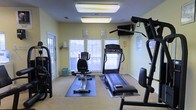 This single-floor fitness room offers a practical and functional space designed to support a well-rounded workout routine. The room is furnished with a variety of exercise equipment, including a treadmill, a recumbent stationary bike, and a multi-function cable weight machine, providing users with both cardiovascular and strength training options. A wall-mounted television adds convenience for entertainment during workouts. The equipment is thoughtfully arranged on protective floor mats over carpeted flooring, maximizing the use of available space while ensuring safety. Natural light filters in through two windows fitted with horizontal blinds, complementing the bright flush-mounted fluorescent ceiling fixtures that keep the room well-lit at all times. The walls are painted a soft, neutral yellow-green, contributing to a calm and energizing atmosphere. Instructional fitness posters are displayed on the walls for reference. A side door provides additional access and ventilation to the space. Overall, this fitness room is a clean, organized, and accessible facility suited for everyday exercise needs.