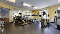 This fitness center is a comprehensively outfitted single-floor gym designed to support a wide range of workout routines. The space is finished with warm yellow walls and durable rubber flooring, creating an energetic yet functional atmosphere throughout. Recessed fluorescent lighting panels illuminate the interior evenly, while multiple windows along the walls invite natural light and offer views of the surrounding greenery, enhancing the overall environment.

The cardio section is well-stocked, featuring multiple treadmills, an elliptical trainer, a stair climber, and a stationary bike, providing ample options for aerobic exercise. A wall-mounted flat-screen television is positioned for easy viewing during workouts. The strength and free-weight area includes a full rack of dumbbells in graduated sizes, a flat bench, and a multi-station cable machine suitable for a variety of resistance exercises. A rowing machine rounds out the equipment selection, adding a full-body cardio and strength option. Floor-to-ceiling mirrors line one wall, visually expanding the space and aiding in proper form during exercise. A glass partition separates an adjoining area, adding a sense of openness. Resistance bands and additional accessories are also available, making this a versatile and practical fitness facility.