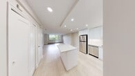 This well-designed 1-bedroom, 1-bathroom unit offers a clean, modern aesthetic throughout its single-floor layout. Light hardwood flooring runs continuously across the open-concept living and kitchen areas, enhancing the sense of space and cohesion.

The kitchen is a standout feature, outfitted with high-gloss white cabinetry extending to the ceiling, a full suite of stainless steel appliances including a refrigerator, electric range, built-in microwave, and dishwasher, and a white waterfall-edge island that doubles as a breakfast bar. Recessed lighting illuminates the space efficiently.

The living room is bright and airy, benefiting from large floor-to-ceiling windows and a glass-panel door that opens directly to an outdoor balcony, offering views of surrounding greenery. Baseboard heating runs along the window walls, ensuring year-round comfort.

The bedroom is a comfortable, well-proportioned space with a sliding glass window, baseboard heating, and light-toned walls. A dedicated closet and an in-unit stacked washer and dryer provide practical storage and laundry convenience.

The bathroom features a clean white tile surround with a soaking tub and shower combination. Throughout the unit, the consistent use of white tones, natural light, and modern finishes creates a calm, contemporary atmosphere.
