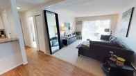 This single-floor residence offers a thoughtfully arranged living space that combines functional design with a bright, welcoming ambiance. The entryway opens directly into the main living area, creating an immediate sense of openness. A striking full-length mirror with a bold black frame serves as a prominent design element, visually expanding the space while adding a contemporary touch.

The living area features a transition from hardwood flooring near the entry to plush carpeting, defining the lounge zone where a dark sectional sofa anchors the room. A light-toned area rug adds warmth and texture underfoot. Natural light flows generously through large sliding glass doors fitted with sheer white curtains, offering a view of an outdoor space beyond and creating an airy atmosphere throughout.

A sleek console or sideboard in dark tones provides practical storage and display space, accented by table lamps that contribute warm ambient lighting. A wall-mounted air conditioning unit ensures year-round comfort. White walls and white panel doors maintain a clean, neutral backdrop, while a hallway visible from the entry suggests additional rooms beyond, contributing to the overall sense of a well-organized, comfortable single-level home.