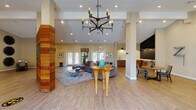 This single-floor community clubhouse offers a thoughtfully designed layout that blends functionality with contemporary style. Guests are welcomed through a bright entryway featuring large glass double doors framed by sidelights and a transom window, flooding the foyer with natural light and views of palm trees beyond.

The expansive main lobby and lounge area commands attention with soaring vaulted ceilings, recessed lighting, and a striking multi-arm industrial chandelier. A distinctive wood-clad structural column serves as a bold focal point, complemented by light wood-look tile flooring throughout. A front reception desk anchors one side of the space, while a generously sized seating area with plush gray sofas, colorful accent pillows, and eclectic sculptural décor creates an inviting atmosphere. A wall-mounted flat-screen TV is integrated into the wood-paneled feature wall.

The kitchenette area is outfitted with warm wood cabinetry, white quartz countertops, a subway tile backsplash, a commercial coffee machine, microwave, beverage refrigerator, and a chalkboard accent wall with counter-height seating.

A dedicated conference room features a large white table surrounded by vibrant orange chairs beneath a rectangular pendant light, with bold abstract artwork adorning the walls. Adjacent to it, a business center provides individual workstations with desktop computers. A cozy nook with a chess table and decorative wall art rounds out this well-equipped community space.