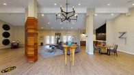 This single-floor community clubhouse offers a thoughtfully designed layout that blends functionality with contemporary style. Guests are welcomed through a bright entryway featuring large glass double doors framed by sidelights and a transom window, flooding the foyer with natural light and views of palm trees beyond.

The expansive main lobby and lounge area commands attention with soaring vaulted ceilings, recessed lighting, and a striking multi-arm industrial chandelier. A distinctive wood-clad structural column serves as a bold focal point, complemented by light wood-look tile flooring throughout. A front reception desk anchors one side of the space, while a generously sized seating area with plush gray sofas, colorful accent pillows, and eclectic sculptural décor creates an inviting atmosphere. A wall-mounted flat-screen TV is integrated into the wood-paneled feature wall.

The kitchenette area is outfitted with warm wood cabinetry, white quartz countertops, a subway tile backsplash, a commercial coffee machine, microwave, beverage refrigerator, and a chalkboard accent wall with counter-height seating.

A dedicated conference room features a large white table surrounded by vibrant orange chairs beneath a rectangular pendant light, with bold abstract artwork adorning the walls. Adjacent to it, a business center provides individual workstations with desktop computers. A cozy nook with a chess table and decorative wall art rounds out this well-equipped community space.
