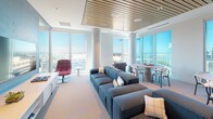 This elegantly designed high-rise lounge occupies a single open floor and is defined by expansive floor-to-ceiling windows on multiple sides, flooding the interior with natural light and offering panoramic views of the surrounding cityscape. The slatted wood ceiling adds warmth and texture, creating a refined contrast against the crisp white walls and soft neutral flooring throughout.

The main lounge area is anchored by a generous modular sectional sofa in slate blue, oriented toward a striking feature wall finished in a deep blue-grey textured surface. Two large flat-screen televisions are mounted side by side above a long, low white media credenza with open shelving and cabinetted storage. A sleek linear fireplace is integrated into a secondary seating zone, accompanied by leather accent chairs and a wall-mounted television, offering an intimate gathering area.

A round dining table with modern chairs is positioned near the windows, making the most of the city views. Additional accent seating, including a bold red swivel chair, provides flexible arrangements throughout the space. Glass-paneled doors lead to an adjacent corridor, maintaining an open and airy feel. The overall ambiance is modern and polished, balancing comfort and sophistication in a light-filled, urban setting.