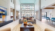 Located at 2332 N 7th St in Phoenix, AZ, this expansive two-story amenity facility offers a sophisticated and thoughtfully designed environment spread across two generous floors. The ground floor commands attention with a dramatic double-height lobby anchored by floor-to-ceiling steel-framed windows that flood the space with natural light and frame sweeping views of a resort-style pool area lined with palm trees. A striking fireplace wall with an integrated large-format display serves as a focal point, flanked by plush lounge seating including boucle armchairs and leather sofas accented with colorful throw pillows. A stylish bar with a dark stone countertop, exposed wood beam ceiling details, and vibrant blue upholstered barstools adds a social hub to the main level. The game and entertainment area features both a shuffleboard table and a billiards table beneath sculptural pendant lighting, complemented by bold contemporary wall art. The upper floor extends the amenity offering with additional lounge nooks, private glass-walled rooms ideal for meetings or quiet retreats, built-in cabinetry with dark stone countertops, and curated artwork throughout the corridors. Neutral-toned patterned carpeting, large-format tile flooring, and a cohesive desert-modern aesthetic tie the entire space together.