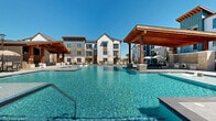 Located at 24720 Morton Ranch Rd in Katy, TX, this outdoor amenity space serves as the centerpiece of a modern residential community. The focal point is a generously sized, resort-style swimming pool with crystal-clear blue water, designed with a multi-section layout that includes both a main swimming area and a separate wading or spa section. The pool deck is finished in large-format, light-toned pavers that extend spaciously around the water's edge, providing ample room for lounging and socializing. Multiple wood-framed pergolas and covered pavilions with ceiling fans are positioned throughout the area, offering shaded gathering and dining spaces. Comfortable chaise lounge chairs, market umbrellas, and outdoor seating are thoughtfully distributed across the deck. The surrounding architecture features a blend of modern farmhouse-style multi-story residential buildings clad in white board-and-batten siding, warm brick, and wood accents with pitched rooflines. The entire space is open to the sky, bathed in natural Texas sunlight, and enclosed by a perimeter fence. The cohesive design, combining rustic wood details with clean contemporary lines, creates an inviting, upscale outdoor environment well-suited for relaxation and community gatherings.