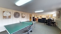 This single-floor space is thoughtfully designed for recreation and relaxation, offering a range of amenities within a cohesive, welcoming layout. The main area is anchored by a full-size ping-pong table set atop neutral carpeting, providing ample room for active play. Vertical wood panel walls painted in soft cream tones give the space a warm, casual character throughout.

The lounge area features a plush blue sofa, a pair of upholstered wingback chairs, and a glass-top coffee table, arranged to encourage comfortable conversation and downtime. Natural light filters in through a row of horizontal windows along one wall, complementing the overhead fluorescent lighting. A large decorative clock and framed wall art add personality and a rustic charm to the space.

Adjacent to the lounge is a dedicated dining area with a dark wood glass-top table and leather-accented seating. The kitchenette is equipped with a double sink set into a granite-style countertop, warm wood cabinetry, a microwave, and a compact refrigerator — providing convenient refreshment options without leaving the space. A decorative octagonal mirror and wall sconce add an elegant touch to this functional nook.

Overall, the space strikes a balance between practicality and comfort, making it well-suited for social gatherings, casual entertaining, or recreational use.