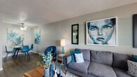 This single-floor residence is styled with a consistent teal, navy, and grey color palette that ties each room together cohesively. The living room serves as the heart of the space, featuring a grey sectional sofa accented with teal and white throw pillows, a live-edge wood coffee table, and a plush area rug. A large sliding glass door fills the room with natural light and provides outdoor access, while a curated gallery wall — anchored by a bold, large-format portrait painting — adds a striking artistic focal point. Textured ceilings are present throughout.

The dining area is compact yet thoughtfully arranged, with a glass-top table and teal chairs positioned beneath botanical-inspired artwork. Adjacent to the dining space, the galley-style kitchen features white cabinetry with brushed nickel hardware, speckled countertops, dark appliances including a black refrigerator and dishwasher, a stainless steel sink, and wood-look flooring that extends throughout the unit.

The bedroom is a standout space, highlighted by a dramatic, oversized geometric starburst headboard that commands the room. A window with sheer curtains allows soft natural light, and coordinating abstract artwork completes the sophisticated look. The bathroom includes a full tub-shower combination with a decorative ombre curtain, wood-look flooring, and clean white tile.