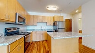 This single-floor residence offers a thoughtfully designed layout that balances functionality and comfort. The kitchen is a standout feature, showcasing light maple cabinetry, granite countertops, and a full suite of stainless steel appliances including a refrigerator, electric range, and built-in microwave. A central island provides additional prep space and serves as a natural gathering point, while warm hardwood flooring ties the kitchen seamlessly into the adjacent living area. The living space is bright and open, benefiting from large windows and a glass door that leads to a private balcony, inviting natural light and outdoor views throughout. The bedroom is carpeted for comfort and features a generously sized closet with wire shelving, offering ample storage. The bathroom is clean and functional, appointed with a wood-toned vanity, white countertop, and a full-width mirror. Neutral wall tones throughout the residence create a calm, versatile backdrop suitable for a variety of interior styles. The overall layout is efficient and well-connected, making excellent use of the available space across every room.