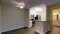 This single-floor space features an open-concept design that seamlessly connects the living area to the kitchen, creating a functional and cohesive layout. The living area is finished with rich, dark hardwood-style flooring and neutral beige walls, contributing to a warm and inviting atmosphere. A ceiling fan with a light fixture is centrally mounted, providing both ventilation and ambient lighting.

The kitchen is thoughtfully designed with crisp white cabinetry that extends to the upper walls, offering ample storage space. Stainless steel appliances, including a gas range with oven and a full-size refrigerator, provide a modern, polished look. A peninsula with a built-in sink and countertop overhang serves as both a prep area and casual dining space, effectively separating the kitchen from the living area while maintaining an open feel. The kitchen area transitions to lighter flooring, visually distinguishing it from the living space. A textured ceiling with flush lighting in the kitchen ensures even illumination throughout. The overall design balances practicality with a clean, contemporary aesthetic.
