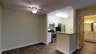 This single-floor space features an open-concept design that seamlessly connects the living area to the kitchen, creating a functional and cohesive layout. The living area is finished with rich, dark hardwood-style flooring and neutral beige walls, contributing to a warm and inviting atmosphere. A ceiling fan with a light fixture is centrally mounted, providing both ventilation and ambient lighting.

The kitchen is thoughtfully designed with crisp white cabinetry that extends to the upper walls, offering ample storage space. Stainless steel appliances, including a gas range with oven and a full-size refrigerator, provide a modern, polished look. A peninsula with a built-in sink and countertop overhang serves as both a prep area and casual dining space, effectively separating the kitchen from the living area while maintaining an open feel. The kitchen area transitions to lighter flooring, visually distinguishing it from the living space. A textured ceiling with flush lighting in the kitchen ensures even illumination throughout. The overall design balances practicality with a clean, contemporary aesthetic.