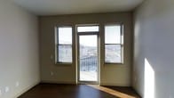 This single-floor space offers a clean, modern layout designed to maximize comfort and natural light. The main living area is characterized by neutral taupe-toned walls and rich dark hardwood flooring, creating a warm yet contemporary ambiance. A trio of windows, complete with adjustable horizontal blinds, flanks a central glass-paned door that opens directly onto a private balcony. This generous fenestration floods the interior with sunlight throughout the day and frames sweeping views of the surrounding cityscape and rolling hills beyond. The balcony, framed by a classic metal railing, extends the living area outdoors and provides an elevated vantage point over the urban landscape. The room's clean lines, uncluttered walls, and open floor plan lend the space a sense of airy spaciousness. Subtle architectural details — including recessed wall plates and a smooth white ceiling — contribute to the overall minimalist aesthetic. The combination of abundant natural light, outdoor access, and panoramic views makes this space particularly well-suited for those who value both a bright interior environment and a strong visual connection to the city surroundings.