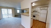 This single-floor residence offers a practical and functional layout designed to maximize the use of space throughout. The kitchen is a standout feature, equipped with white cabinetry, a white electric range, and a built-in overhead range hood. Hardwood flooring adds warmth and durability to the kitchen area, while a pass-through window with a countertop ledge connects it directly to the adjacent living space, promoting an open and communicative flow between the two areas. The living room is generously sized, featuring wall-to-wall carpet and multiple windows that allow natural light to fill the room, creating a bright and welcoming atmosphere. A door in the living area provides direct access to the outdoors. The textured ceilings are consistent throughout, contributing to the home's classic character. The open-concept connection between the kitchen and living room makes the space feel cohesive and well-suited for both everyday living and entertaining. Overall, the layout is efficient and straightforward, with each area clearly defined while remaining visually connected to the rest of the home.