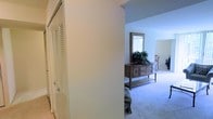 This single-floor residence offers a comfortable and well-organized layout designed for easy living. The interior is finished with soft wall-to-wall carpeting throughout, lending a warm and quiet feel to the space. A hallway connects the living areas, lined with louvered bifold closet doors that provide ample built-in storage while maintaining a clean, classic aesthetic. The living and dining area is open and airy, bathed in natural light that streams in through large windows dressed with vertical blinds. A wooden credenza with decorative diamond-panel detailing serves as a functional accent piece, topped with lush greenery and a framed mirror that enhances the sense of depth and brightness in the room. Neutral wall tones in soft white and light grey create a calm, cohesive palette throughout. The overall ambiance of the space is relaxed and inviting, with a traditional design sensibility that balances practicality and comfort. The single-level layout ensures effortless flow between rooms, making the space accessible and easy to navigate.