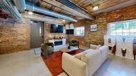 Located at 250 S Depot St in Pendleton, SC, this single-floor space showcases a compelling blend of historic industrial character and contemporary design. The open-concept layout seamlessly connects the kitchen and living areas, making efficient use of its compact footprint.

The living area is anchored by striking exposed brick walls that lend warmth and texture throughout. A reclaimed wood plank ceiling with visible steel beams and exposed ductwork reinforces the industrial aesthetic, while track lighting illuminates the space effectively. Polished concrete floors provide a clean, durable foundation that complements the raw architectural details. A wall-mounted fireplace with a media console serves as a focal point, flanked by modern lounge chairs and a plush sofa atop a rich terracotta area rug. A gridded window allows natural light to filter in, offering views of the greenery outside.

The kitchen features sleek, light-toned cabinetry with black hardware, white countertops, and stainless steel appliances including a large side-by-side refrigerator and dishwasher. A white kitchen island with bar seating provides additional workspace and casual dining. Exposed ductwork and wire cable trays continue the industrial theme overhead, tying both areas together cohesively. The overall ambiance is stylish, functional, and full of character.